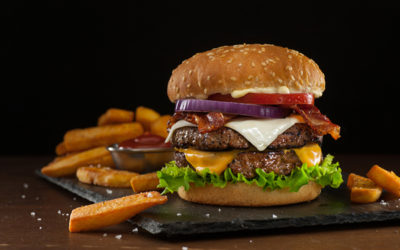 High resolution digital capture of a steakhouse-style double bacon cheeseburger with steak fries. This cheeseburger is made with two patties of ground steak, Cheddar and Monterey Jack cheeses, crispy bacon slices, fresh tomatoes, lettuce, and onion, all on a sesame seed bun. A ramekin of ketchup is visible amongst the fries. Background is dark and atmospheric, with room to expand and place copy.